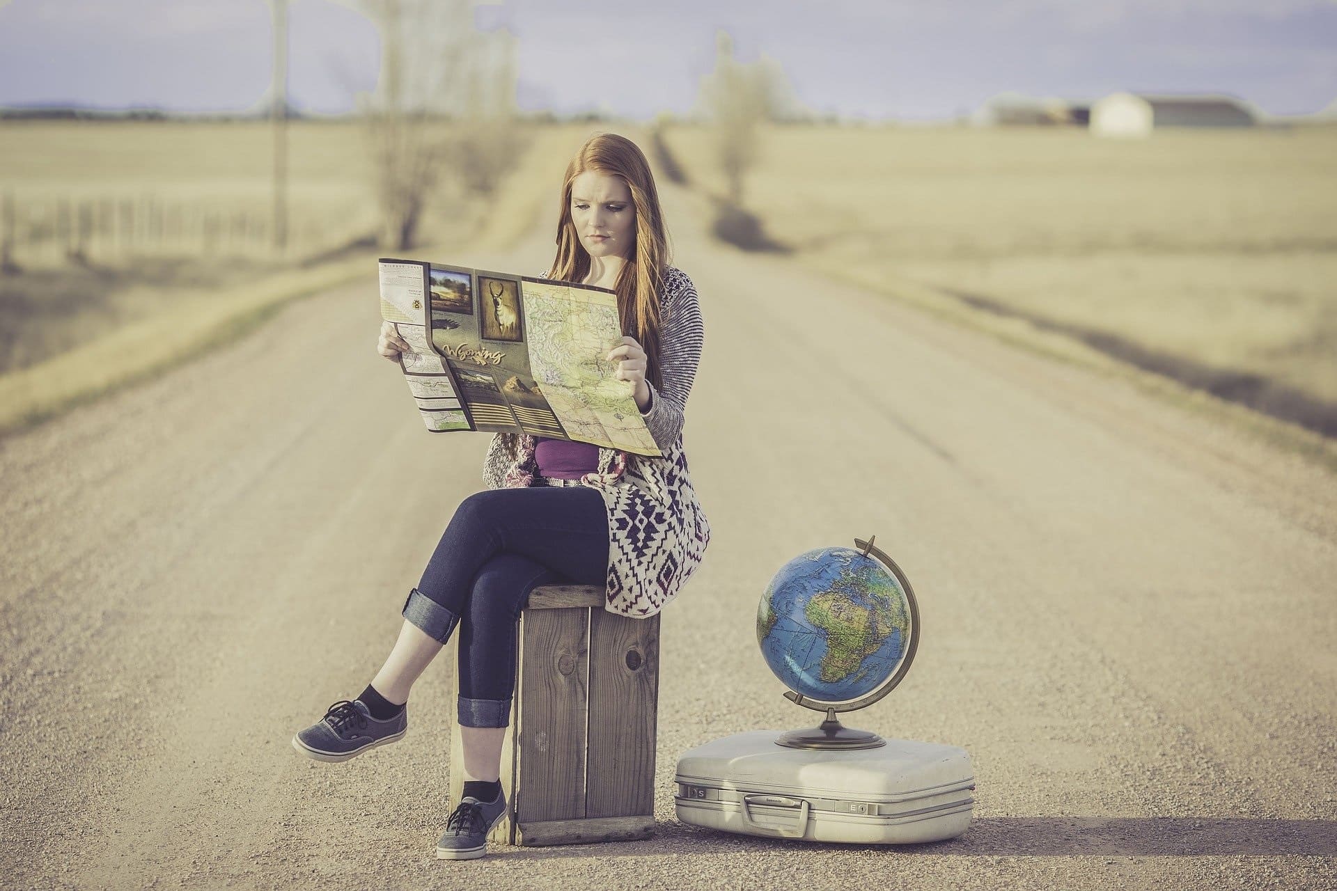exigences douanières femme assise sur une valise au milieu d'une route en train de lire le journal un globe terrestre posé à côté d'elle