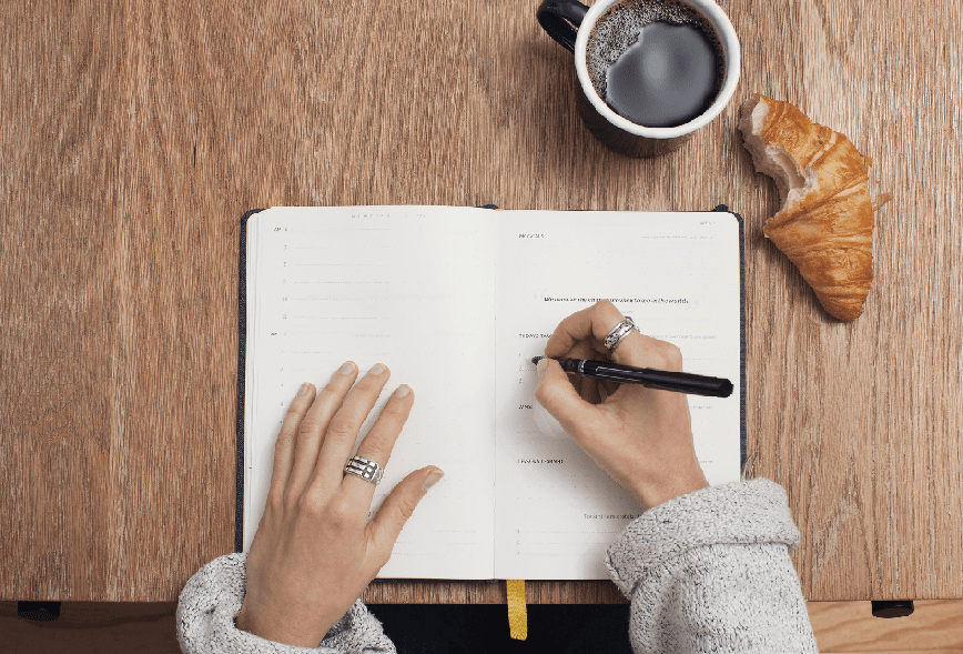 femme expatriée écrivant au stylo dans un cahier de notes ouvert posé sur une table pour préparer son retour en France, un café et un croissant au beurre posés sur la table