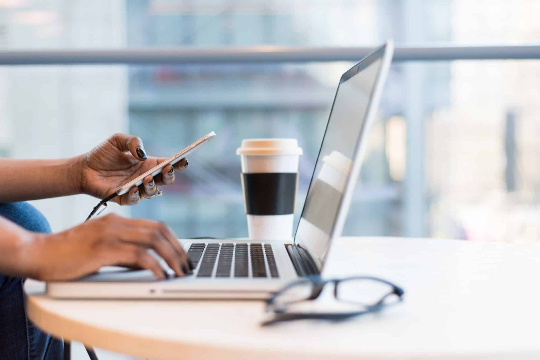femme tapant sur le clavier de son ordinateur portable avec son téléphone dans sa main gauche, lunettes de vue et tasse de café posés sur la table